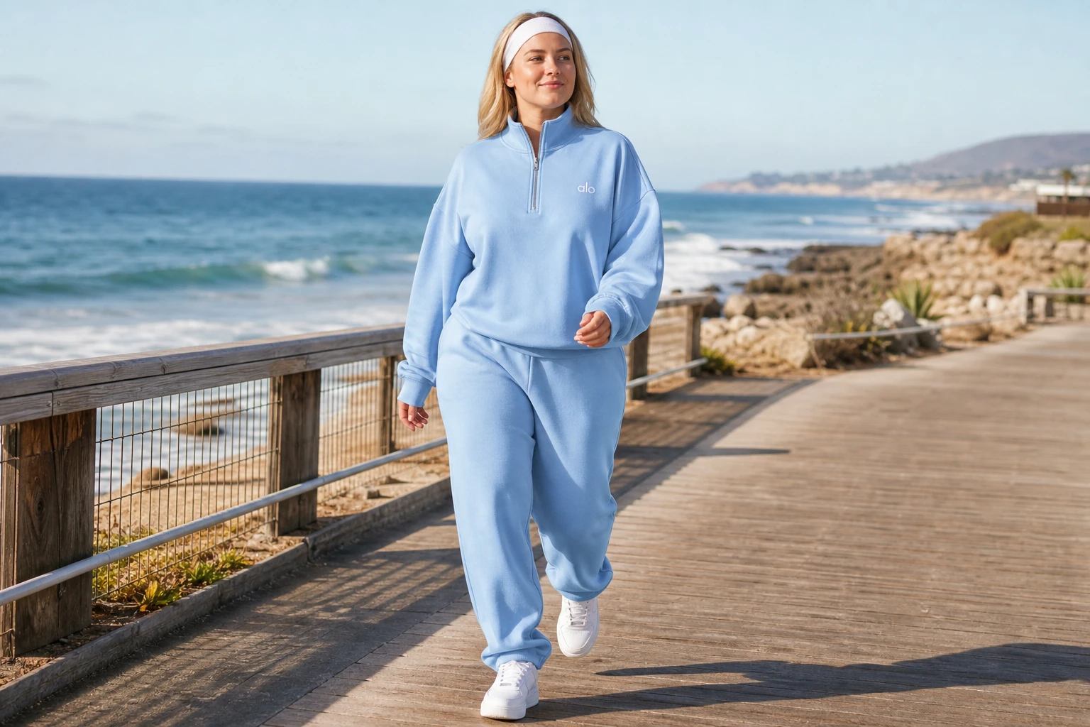 Model in a blue quarter-zip pullover walking a coastal boardwalk