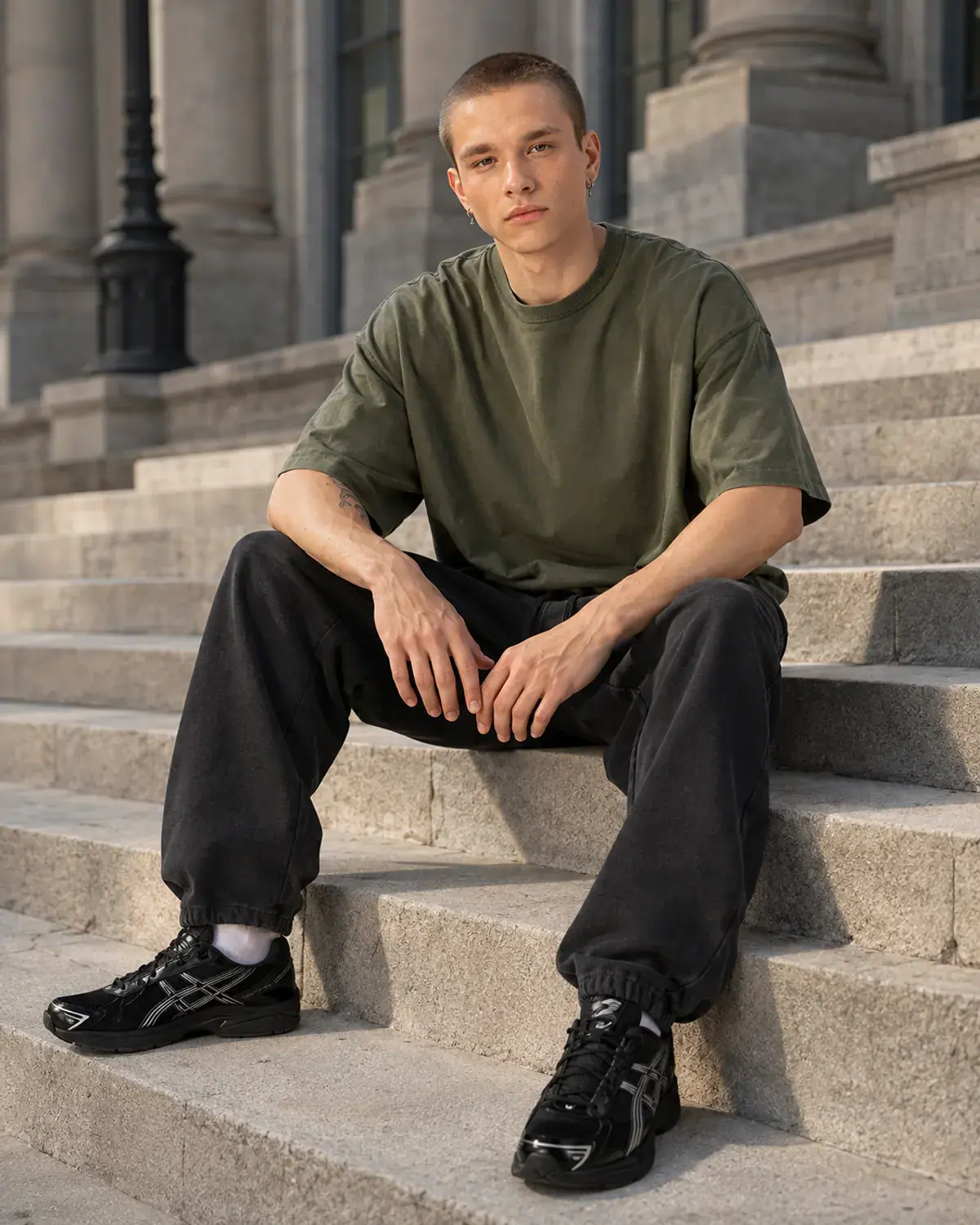 Man in an olive baggy crop tee seated on stone civic steps