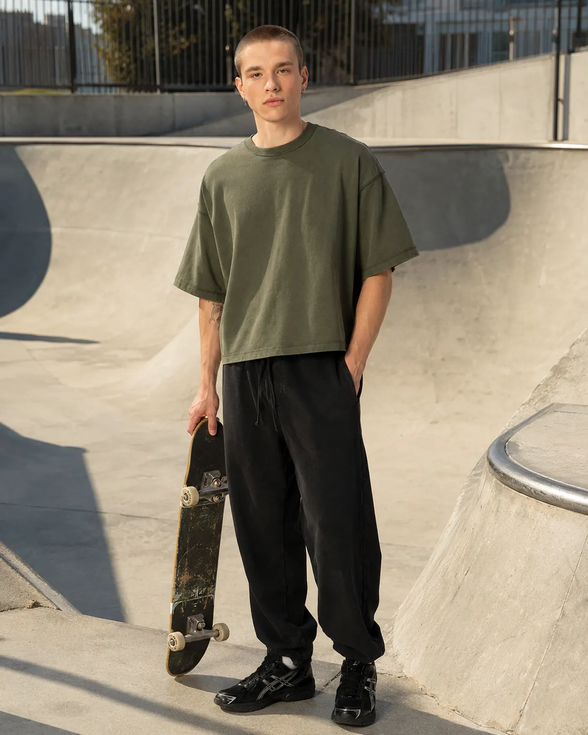 Man in an olive baggy crop tee holding a skateboard at a concrete skatepark