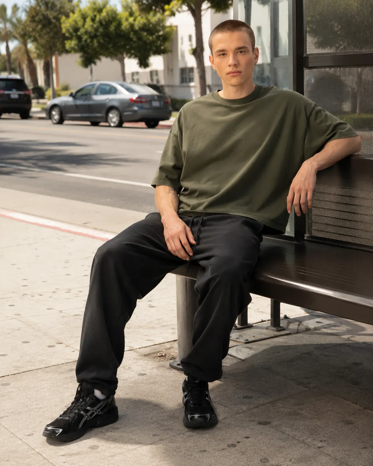 Man in an olive baggy crop tee seated at a palm-lined bus stop
