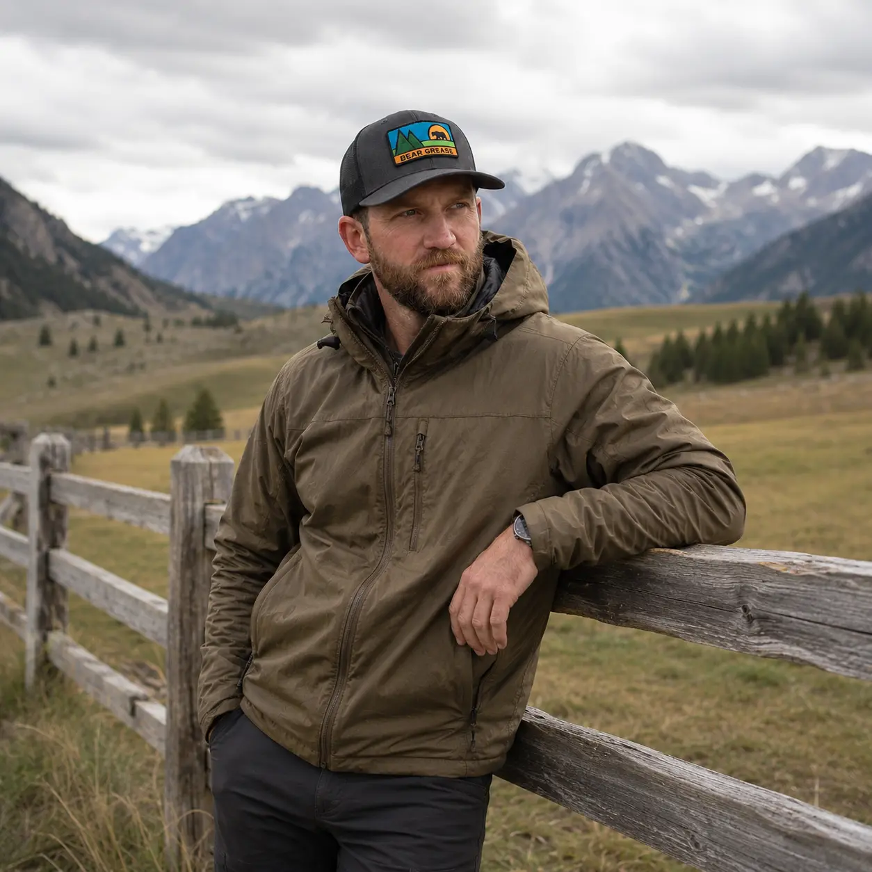 Model leaning on a weathered ranch fence with snowy peaks behind, wearing the Bear Grease trucker hat
