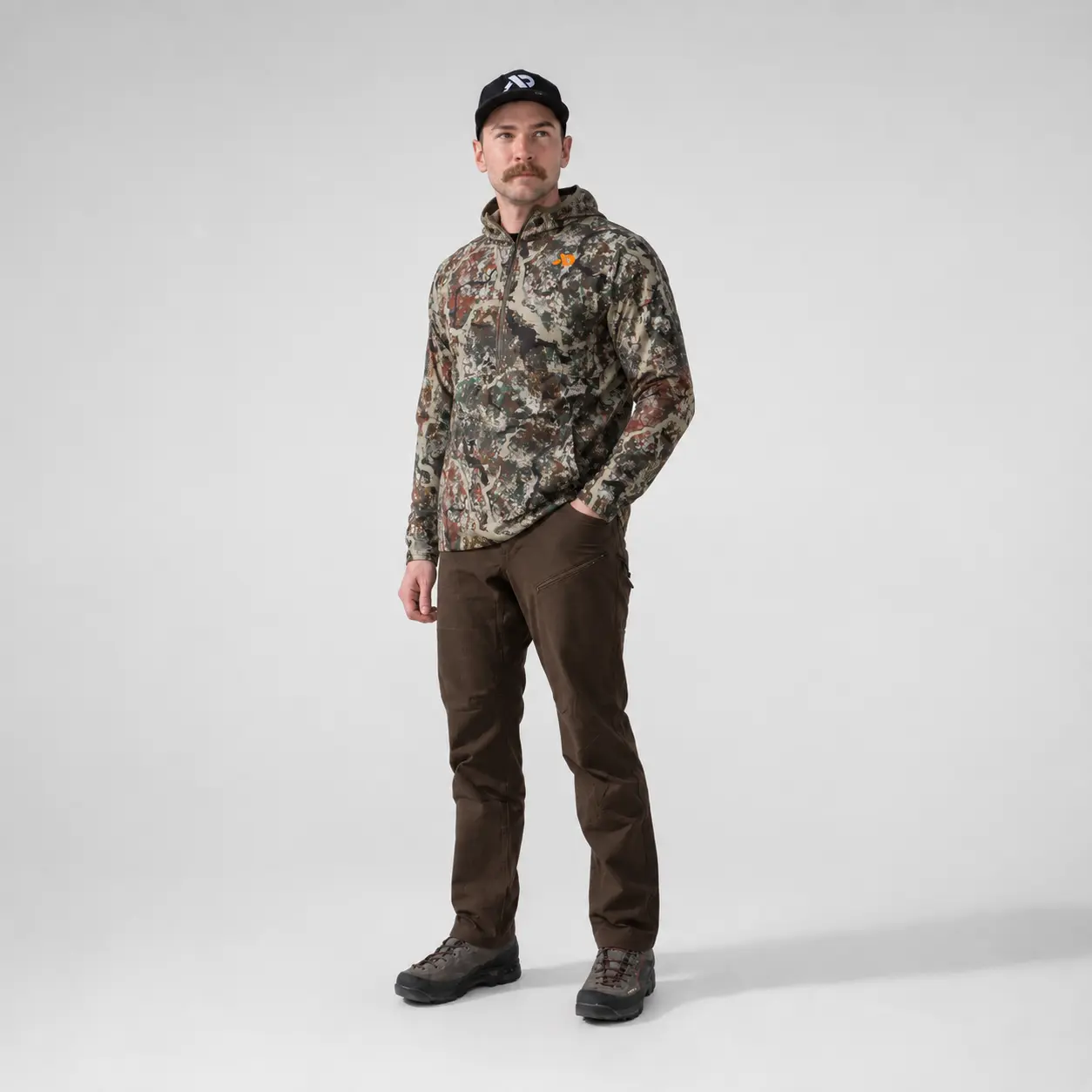 Full length studio shot of a model wearing the Navigator Hoody in First Lite camo with brown pants