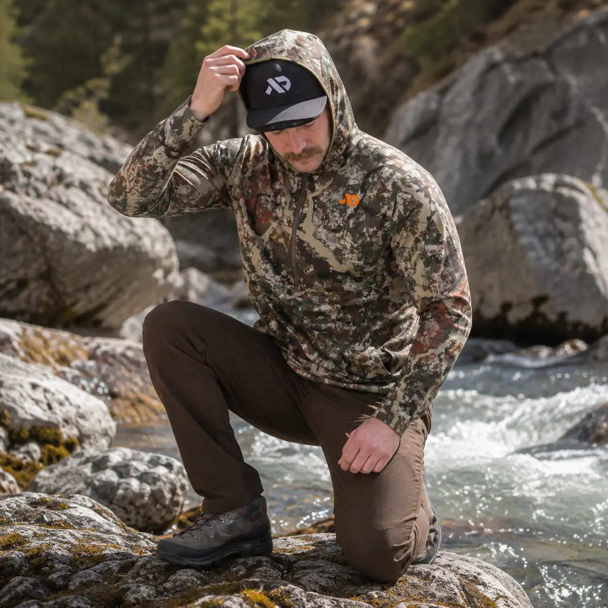 Model kneeling on rocks beside a mountain creek, pulling the Navigator Hoody hood over a cap